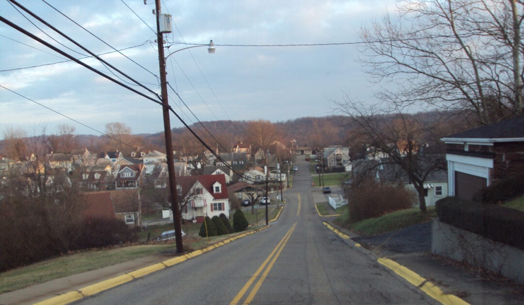 A residential street slopes downhill, lined with houses, leafless trees, power lines, and yellow curbs; the sky is partly cloudy and distant hills are visible in the background.