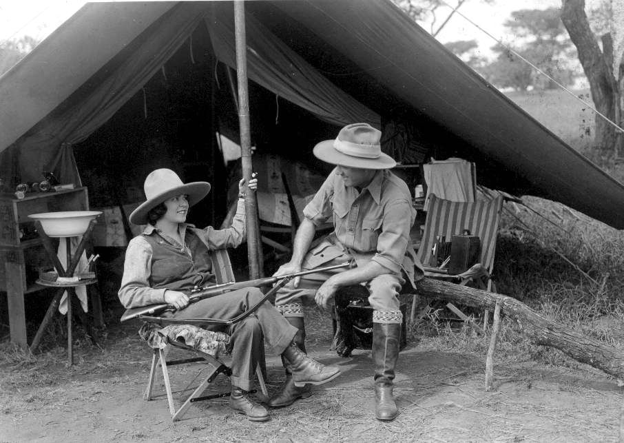 A woman and a man in wide-brimmed hats sit in front of a canvas tent. The woman, in boots and trousers, holds a rifle while smiling at the man, who is dressed in safari gear and sitting beside her. Outdoor supplies are visible behind them.