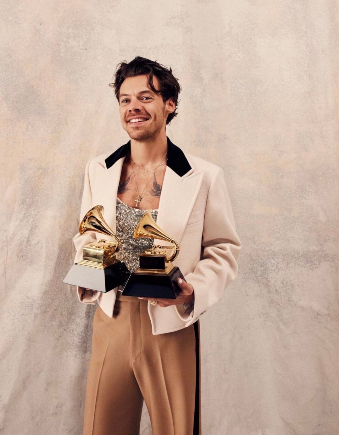 A smiling man wearing a cream-colored jacket and gold sequin top holds two Grammy awards in front of a neutral, textured backdrop.