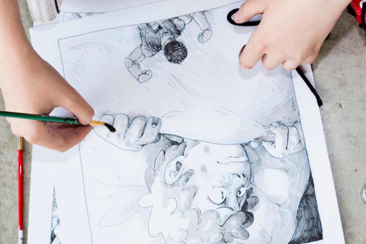 A person wearing red shoes and carrot-patterned pants is sitting on the floor, drawing and coloring a black-and-white comic illustration with a paintbrush. The comic pages are spread out on the ground.