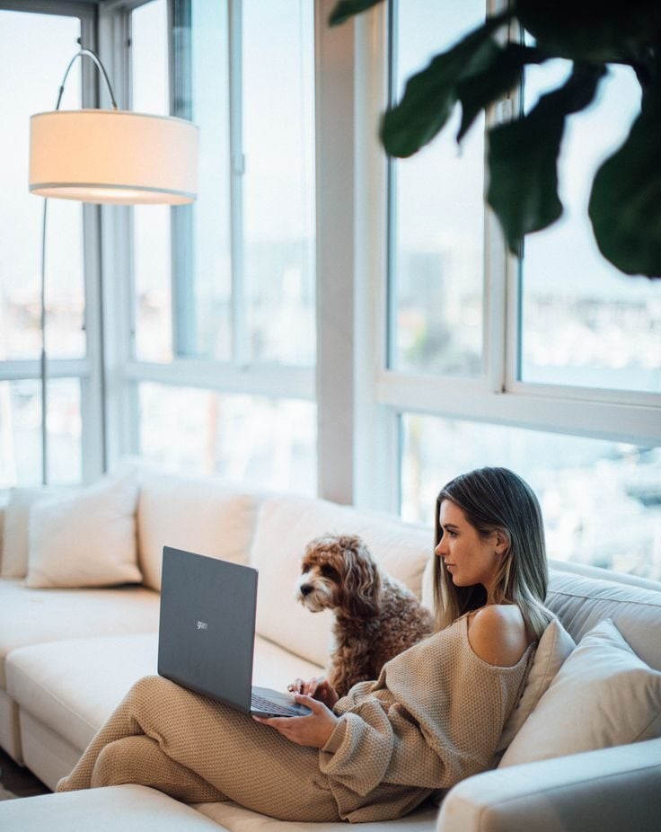A woman in a cozy beige outfit sits on a white sofa with a laptop on her lap. A small brown and white dog sits beside her. Large windows in the background let in natural light, revealing a marina view.