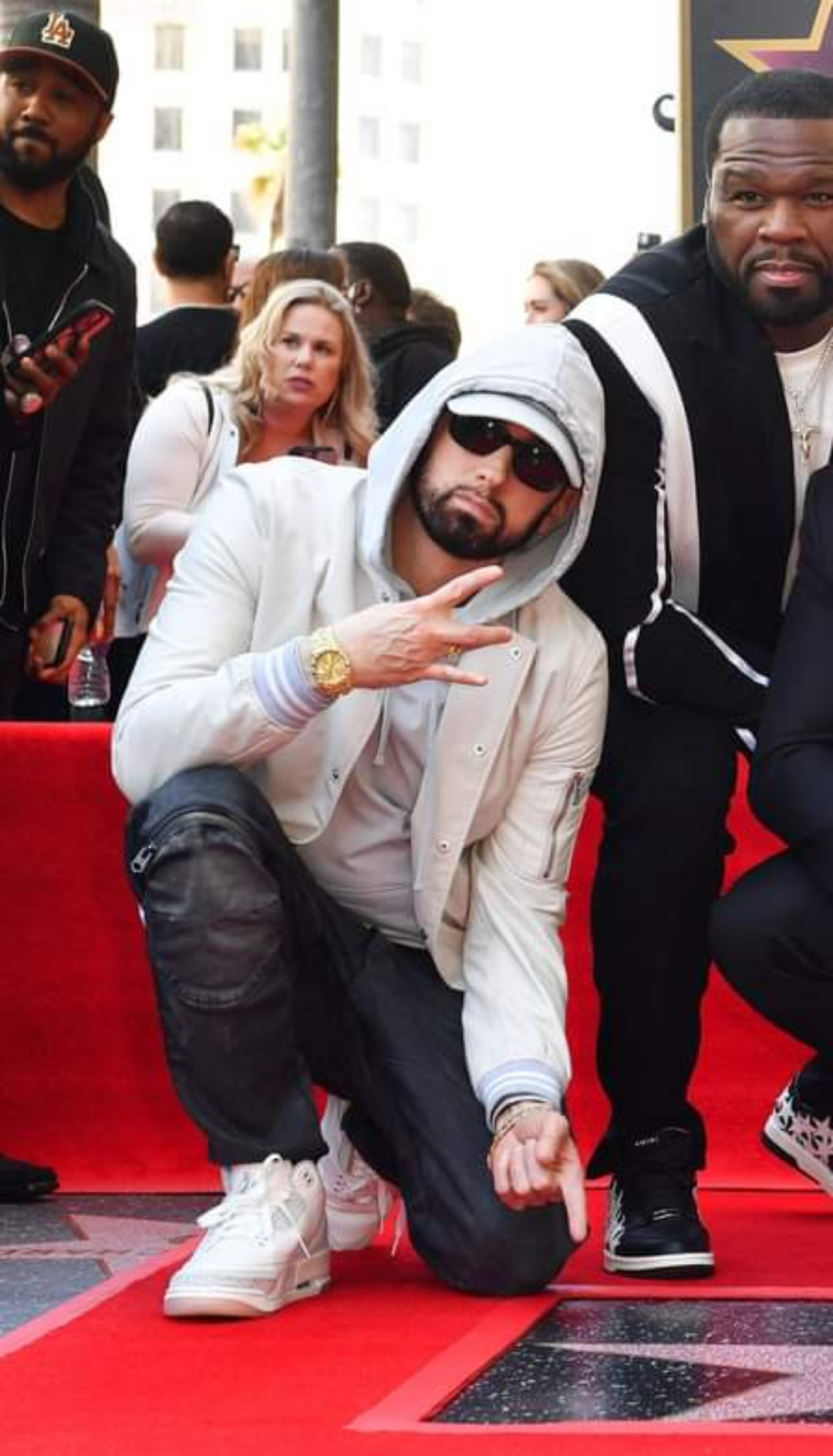 A man in sunglasses, a hoodie, and a light jacket crouches on a red carpet, flashing a peace sign, next to another man in a black and white tracksuit. People stand in the background.