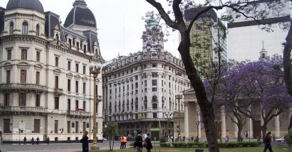 People walk through a city park lined with blooming jacaranda trees, with historic European-style buildings and tall modern structures in the background.