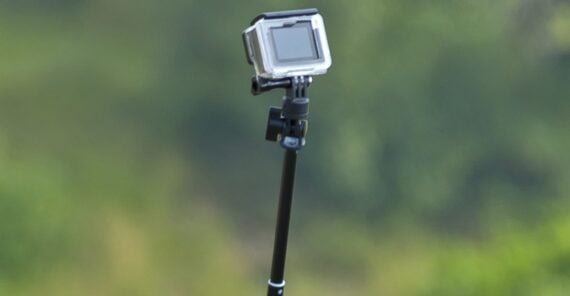 A close-up of an action camera mounted on a black monopod, set against a blurred green outdoor background.