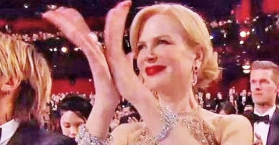 A woman in formal attire with blonde hair and red lipstick is smiling and clapping in an audience at a formal event, surrounded by other well-dressed people.