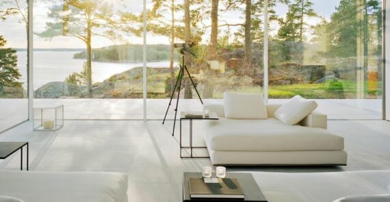 A modern living room with white minimalist furniture and floor-to-ceiling glass walls overlooks a scenic outdoor landscape of trees, rocks, and water. Sunlight streams in, and a telescope faces the view.