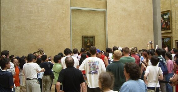 A large crowd of people stands in front of the Mona Lisa painting, protected behind glass, at a museum. Many visitors are taking photos and looking at the artwork. Other framed paintings hang on the walls.