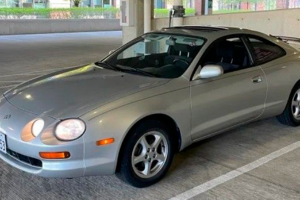 A silver two-door coupe car with pop-up headlights is parked in a covered parking garage with daylight coming through the side.
