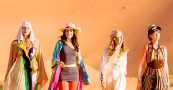 Four women dressed in colorful, fashionable outfits walk confidently in a desert, with sand dunes in the background under a clear sky.