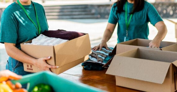 Two people wearing green shirts and lanyards pack folded clothes into cardboard boxes on a wooden table, possibly for donation or charity, with some produce visible in the foreground.
