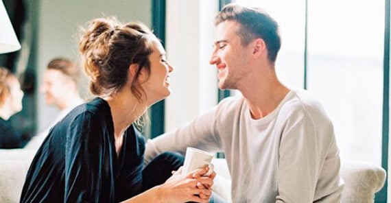 A woman and a man sit close together on a couch, smiling and looking into each other's eyes while holding hands. The woman holds a white mug, and natural light streams in from a window behind them.