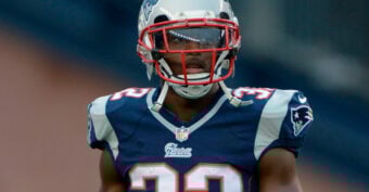 A football player wearing a New England Patriots uniform and helmet with a tinted visor stands on the field. The jersey number is 32.