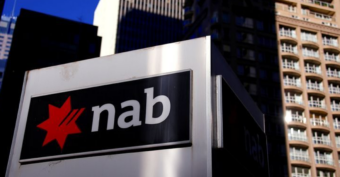 A sign with the NAB logo, featuring a red star and black background, is shown in front of tall city buildings under sunlight.