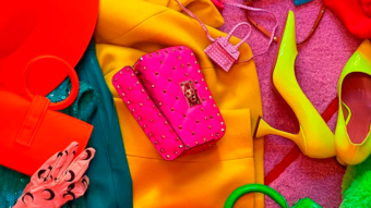 A vibrant flat lay of colorful fashion accessories, including a pink studded handbag, neon green high heels, orange gloves, a yellow coat, a red hat, and other bright items on a pink background.