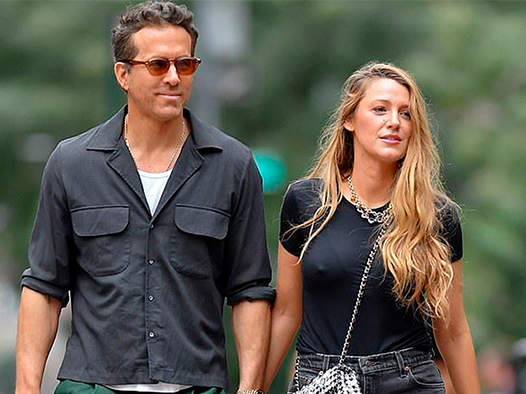 A man in sunglasses and a dark shirt walks outdoors holding hands with a woman in a black t-shirt and long blonde hair; both appear relaxed and are smiling slightly.