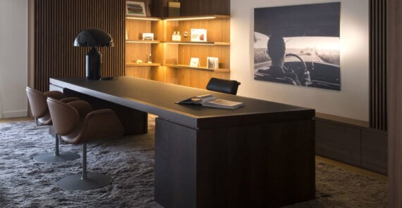A modern office with a long dark desk, three brown chairs, a black lamp, open books, wall shelves with books and decor, and a large black-and-white photo of a person driving on the wall.