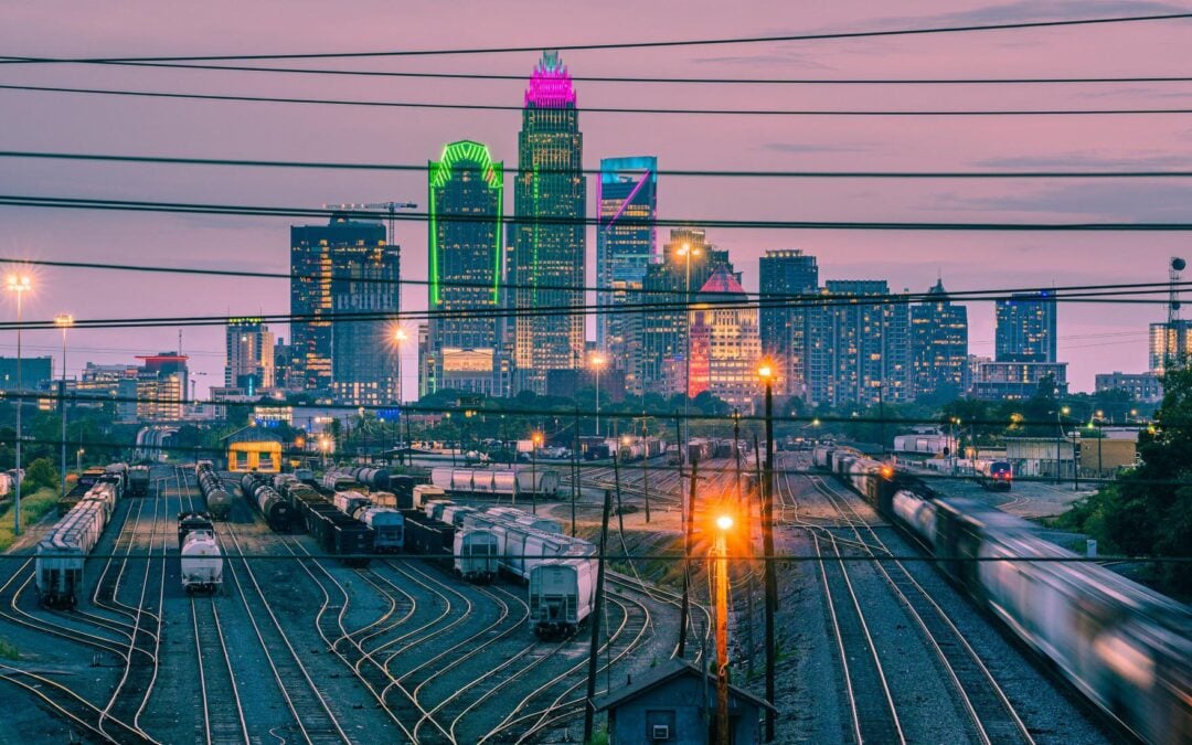 A vibrant city skyline at dusk with colorful, illuminated skyscrapers in the background and multiple train tracks with stationary and moving trains in the foreground, seen through overhead power lines.