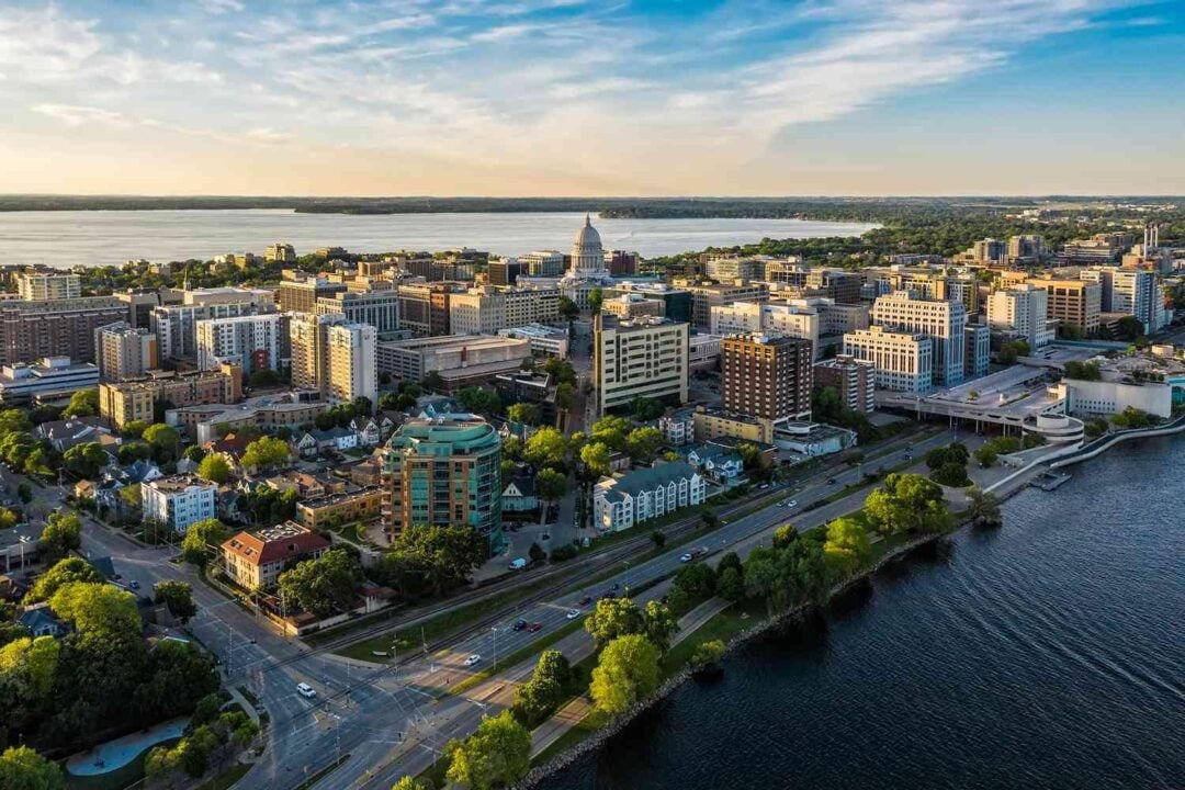 Aerial view of a cityscape with a domed capitol building, surrounded by various buildings, green trees, and bordered by a large lake under a partly cloudy sky.