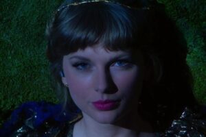 A woman with blonde hair and pink lipstick gazes directly at the camera, lying on a moss-covered surface and partially illuminated by dramatic lighting.
