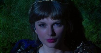 A woman with blonde hair and pink lipstick gazes directly at the camera, lying on a moss-covered surface and partially illuminated by dramatic lighting.