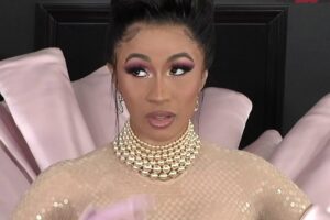 A woman with dramatic makeup and a pearl necklace stands in front of a pink, fan-shaped backdrop, wearing a sheer, nude-colored outfit and long gloves.