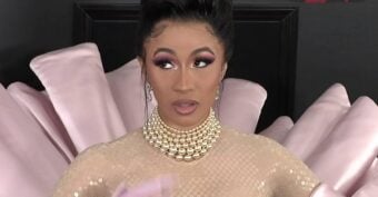 A woman with dramatic makeup and a pearl necklace stands in front of a pink, fan-shaped backdrop, wearing a sheer, nude-colored outfit and long gloves.