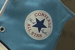 A close-up of a light blue Converse Chuck Taylor All Star sneaker, showing the white circular logo with a blue star and red text on the ankle area. The sneaker has white laces and a white rubber sole.