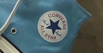 A close-up of a light blue Converse Chuck Taylor All Star sneaker, showing the white circular logo with a blue star and red text on the ankle area. The sneaker has white laces and a white rubber sole.