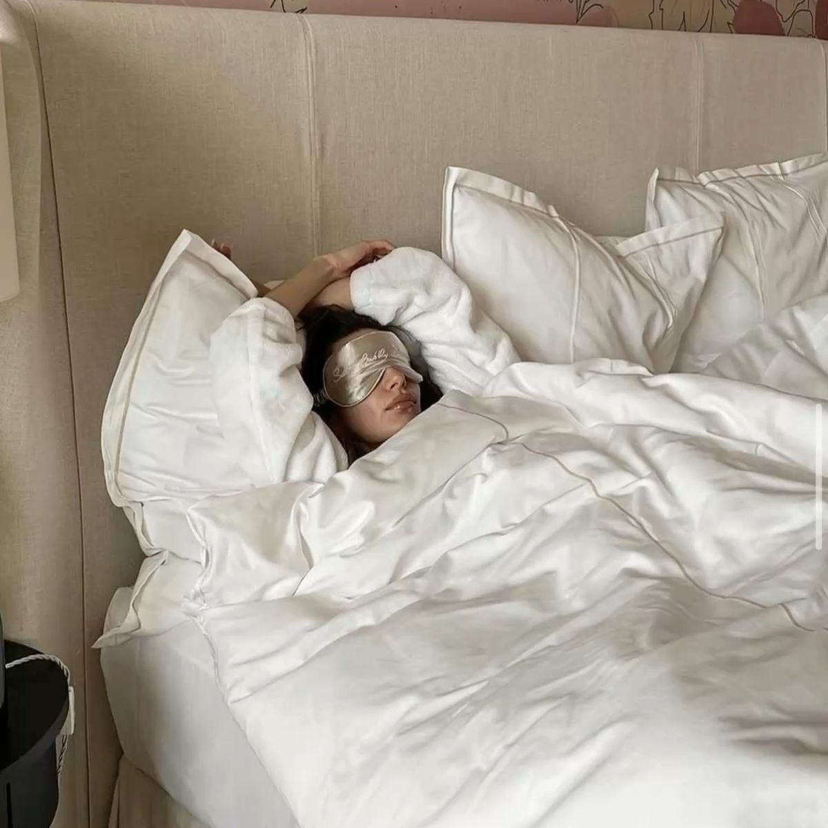 A person wearing a sleep mask and a white robe is lying in bed under white blankets, resting with arms stretched above their head on a large bed with multiple pillows.