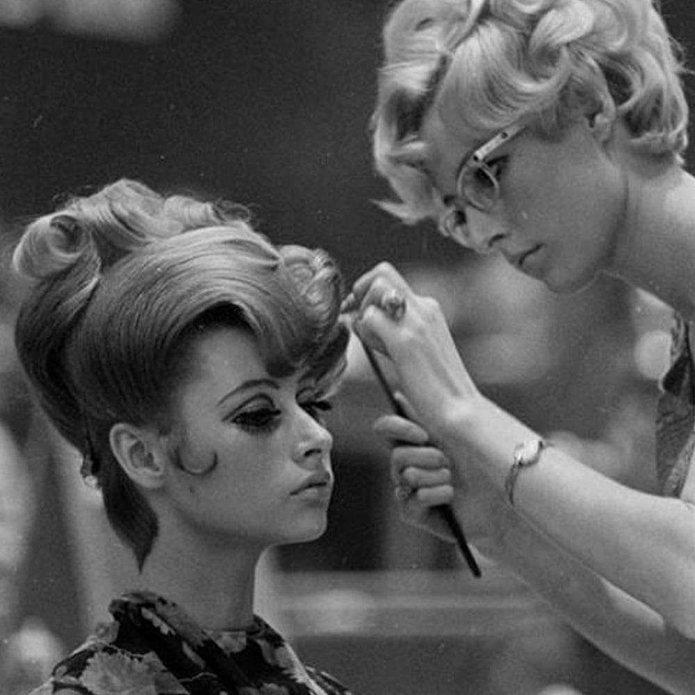 A hairstylist in glasses styles a woman’s voluminous, vintage hairdo with a comb. The seated woman wears dramatic eyeliner and a floral top, showing a classic 1960s fashion and beauty scene.