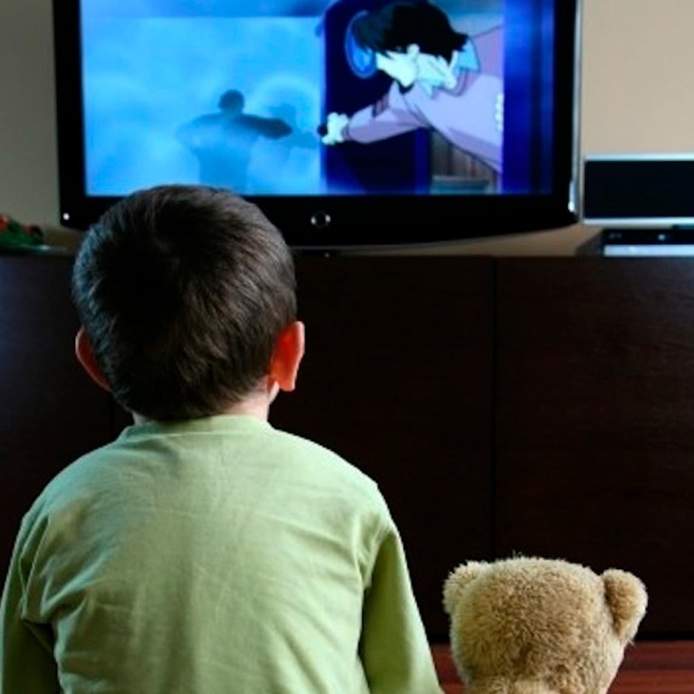 A young child sits on the floor facing a television, watching an animated cartoon. A teddy bear is beside the child, also facing the TV. The scene is set in a cozy living room.