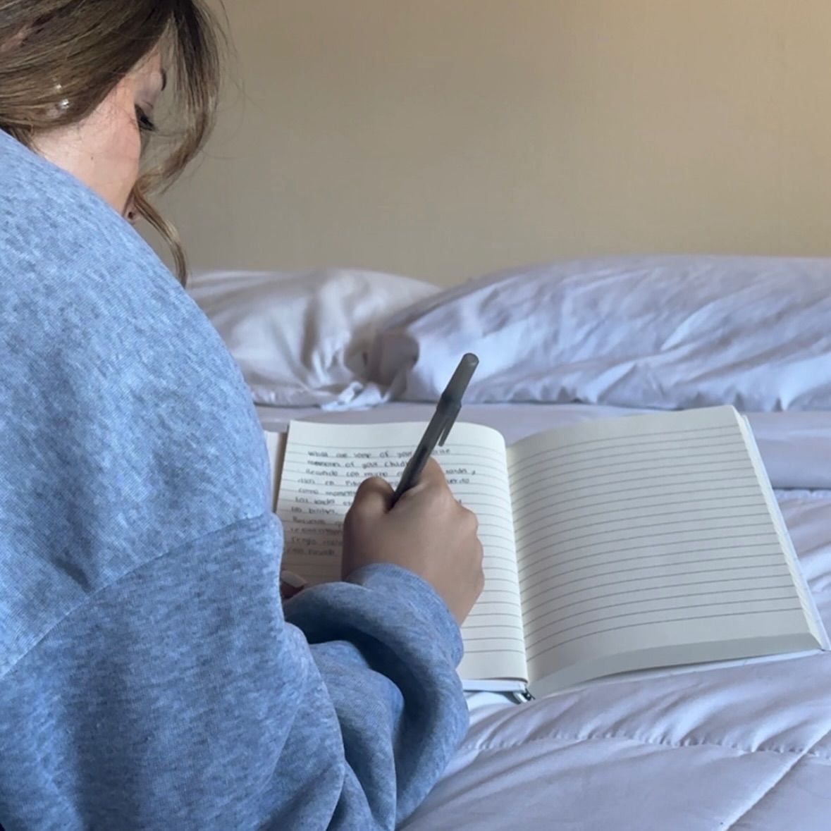 A person in a blue sweatshirt is lying on a bed, writing in a notebook with a pen. The notebook is open, with one page filled with writing and the other page blank.