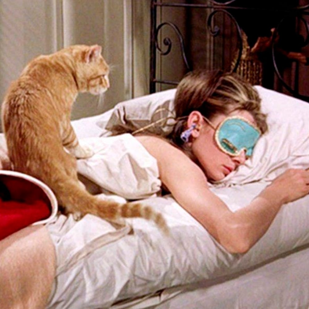 A woman wearing a blue sleep mask lies face down in bed while a ginger cat sits beside her on the blanket, watching her sleep.