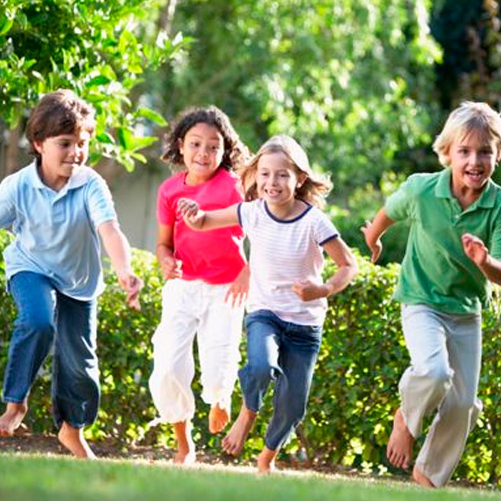 Four children are running barefoot on grass in a garden, smiling and enjoying a sunny day. Green bushes and trees are in the background. The kids appear happy and energetic.