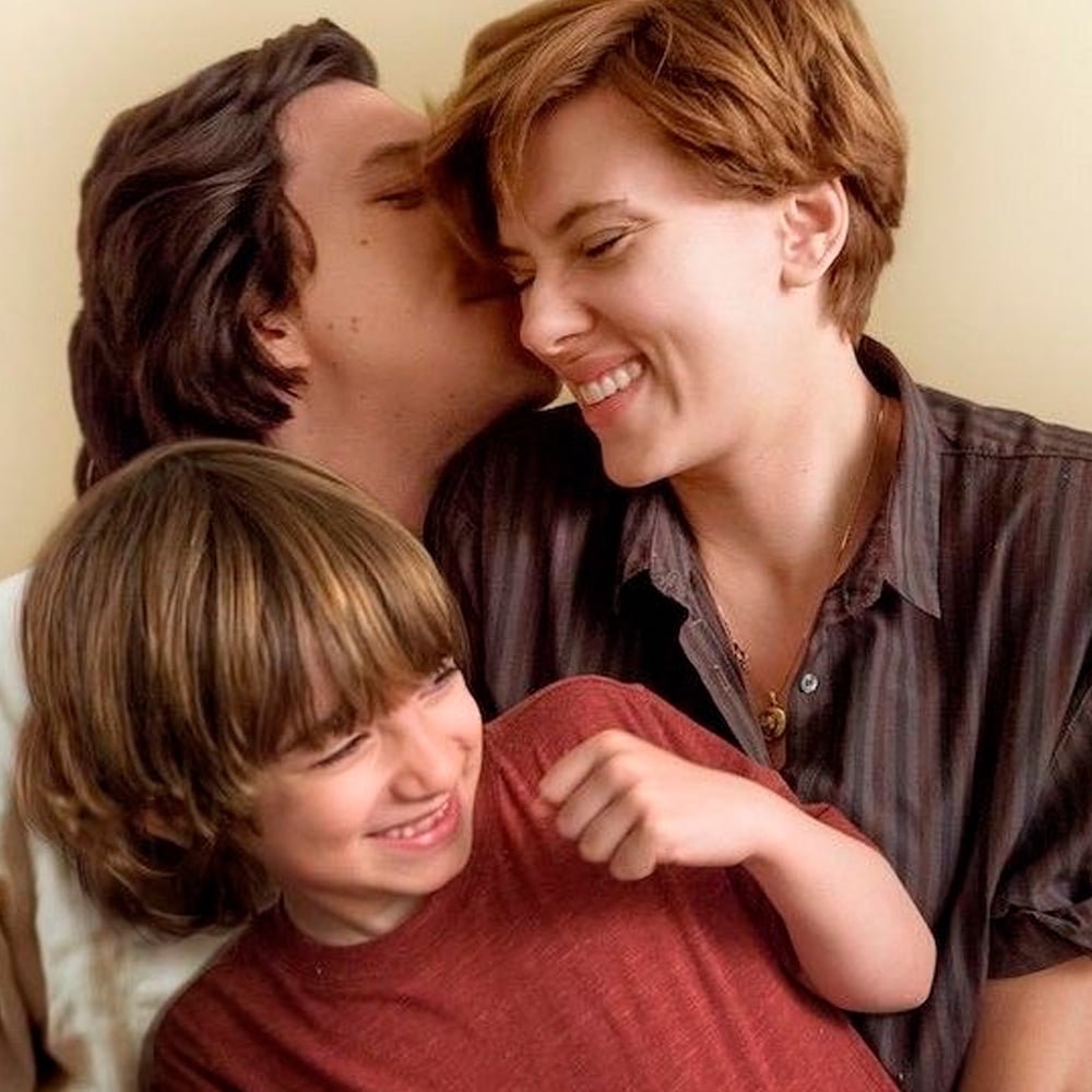 A happy family of three sits closely together, smiling and laughing. The man kisses the woman’s head while she grins, and a young boy in front of them looks joyful, wearing a red shirt.