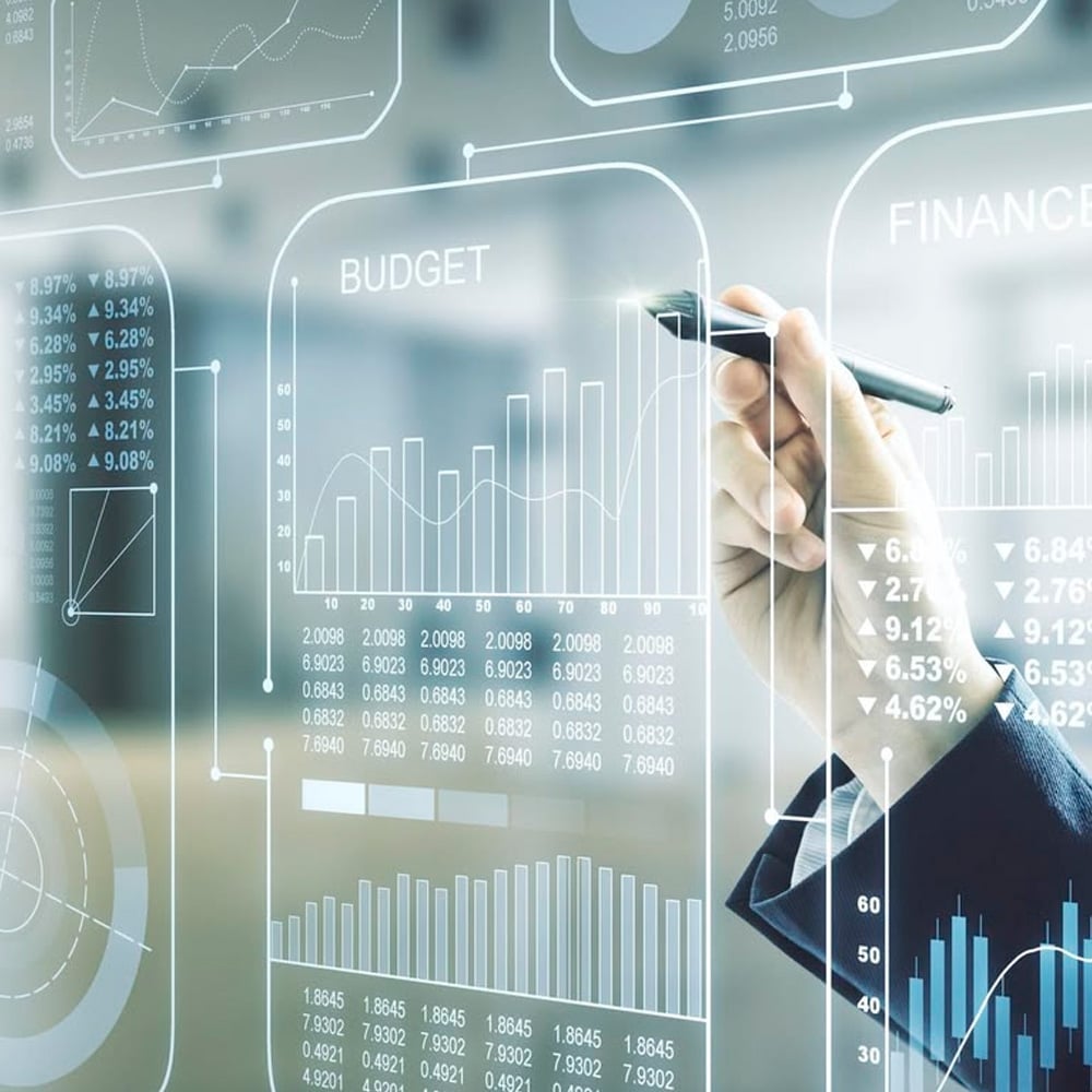 A hand holding a pen points to transparent virtual graphs and charts with financial data, including budget and finance metrics, on a digital interface.