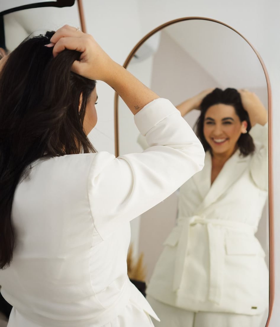 A woman in a white suit smiles while adjusting her hair, looking at herself in a mirror. The room is softly lit and the background is neutral.