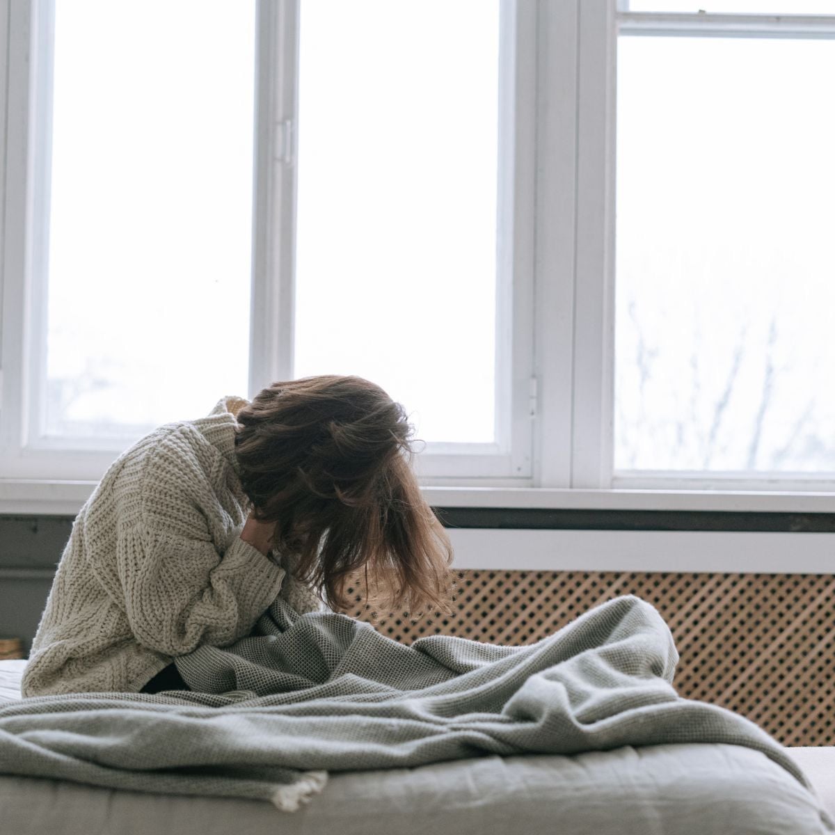 A person sitting on a bed, hunched over with their head down and hands covering their face, wearing a cozy sweater and wrapped in a blanket, in a room with large windows letting in natural light.