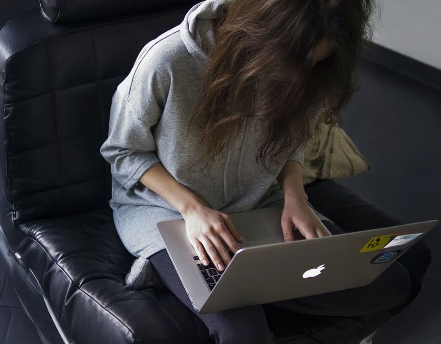 A person with long brown hair, wearing a gray hoodie, sits on a black leather chair and types on a silver MacBook laptop. The person is looking down at the screen, and some stickers are visible on the laptop.
