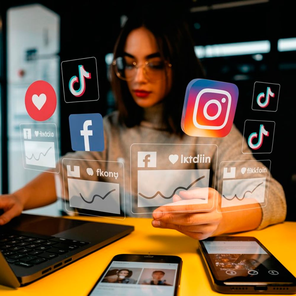 A woman sitting at a desk with a laptop and smartphones interacts with floating social media icons, including Instagram, Facebook, TikTok, and graphs representing digital engagement and analytics.
