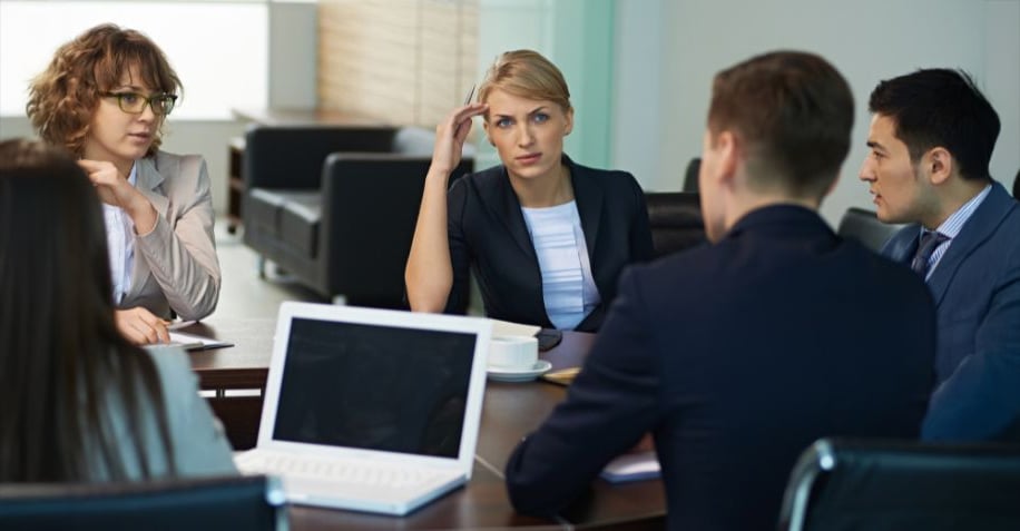 Five people in business attire sit around a table having a serious discussion in an office setting. A laptop is open in the foreground, and one woman looks concerned while listening to others.
