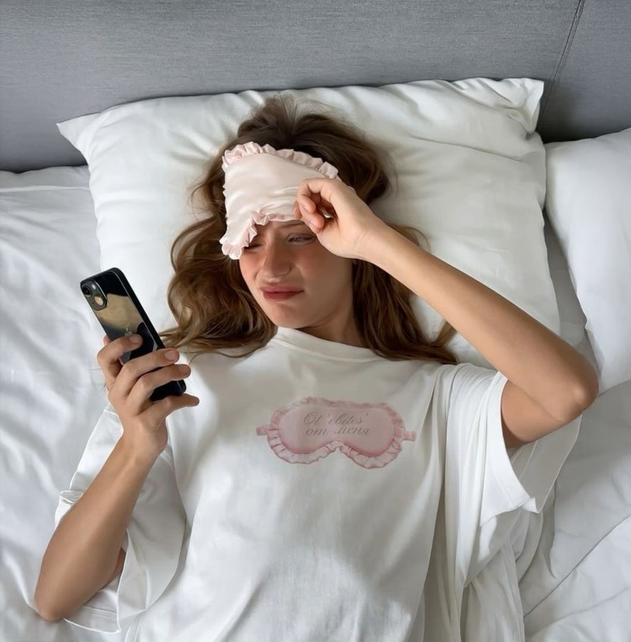 A woman lying in bed with white sheets, wearing a white shirt and a pink sleep mask pushed up on her forehead, looks at her phone and smiles slightly.