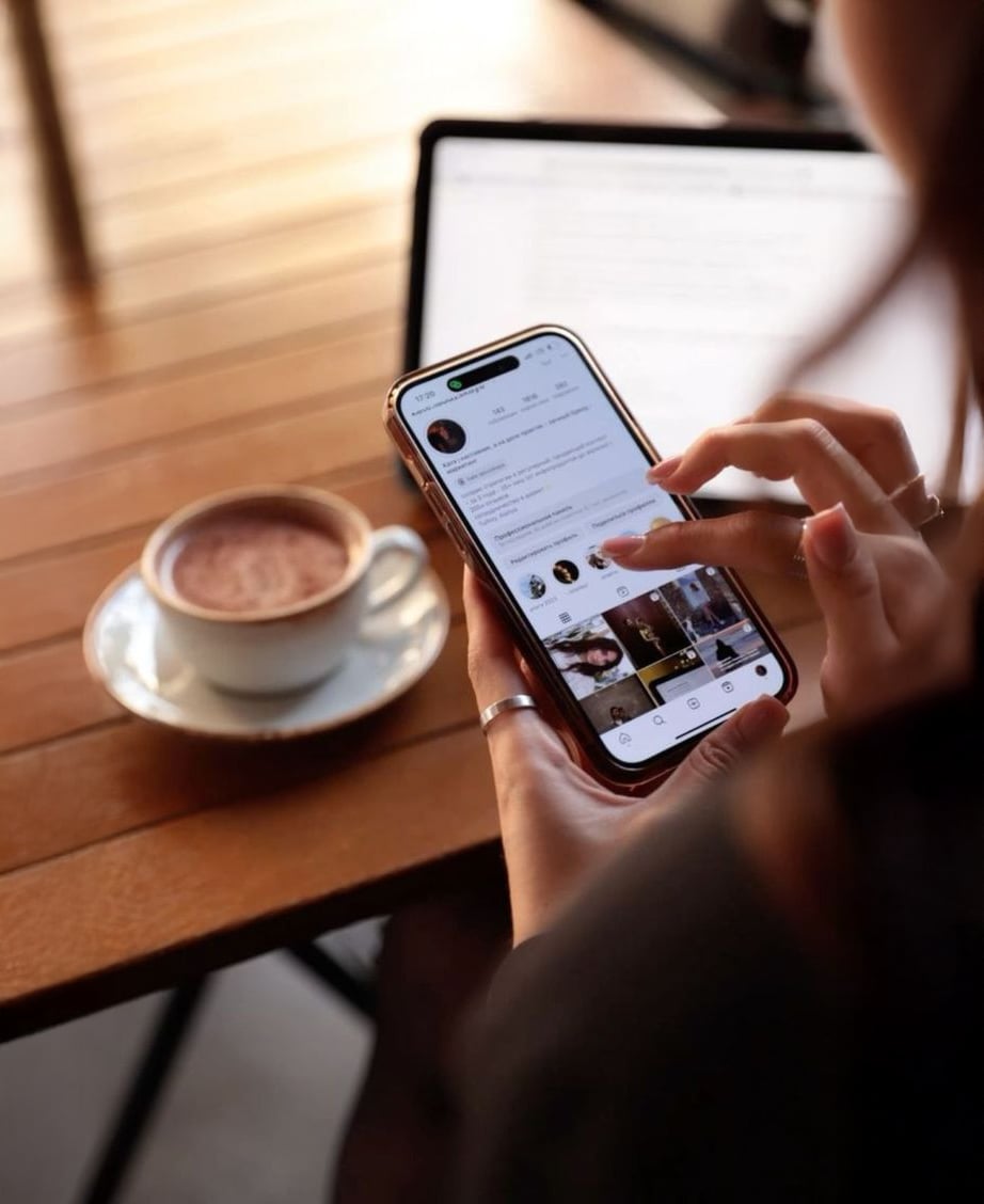 A person uses a smartphone to browse social media at a wooden table, with a cup of hot chocolate and an open laptop in the background.