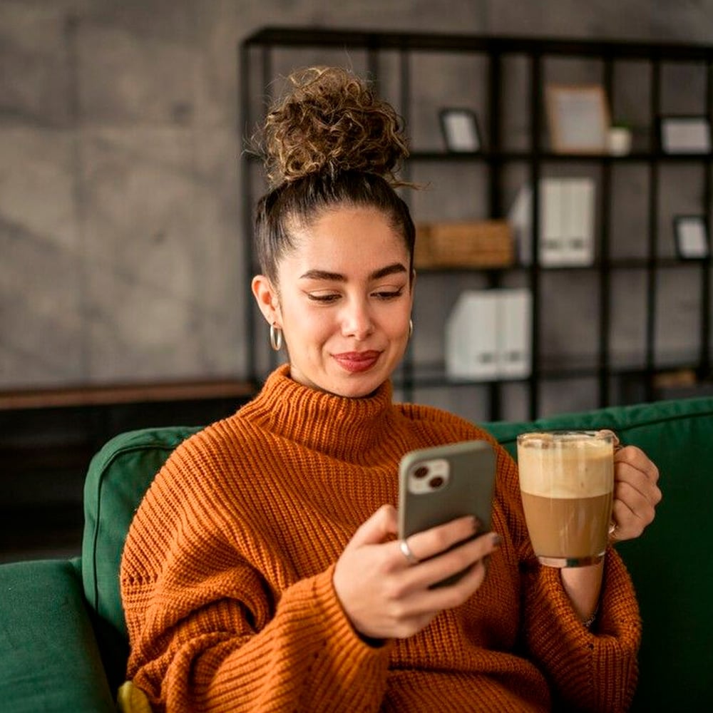 A woman with curly hair in a bun, wearing a brown sweater, sits on a green couch holding a mug of coffee in one hand and looking at her smartphone with a smile. There is a shelf in the background.