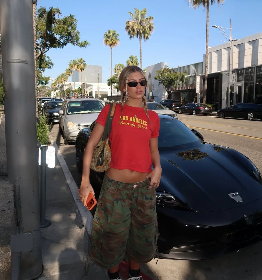 A woman wearing a red "Los Angeles Beverly Hills" t-shirt, camo baggy pants, and sunglasses stands on a sunny street lined with palm trees and parked cars, holding a phone and a gold handbag.