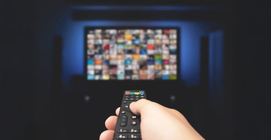A hand holding a TV remote is pointed at a large television screen displaying a grid of colorful, blurred thumbnails, suggesting streaming or browsing for shows or movies in a dark room.