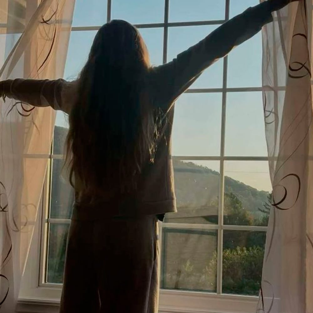 A person with long hair stands by a large window, arms outstretched, opening sheer curtains to let in morning sunlight with a view of trees and hills outside.