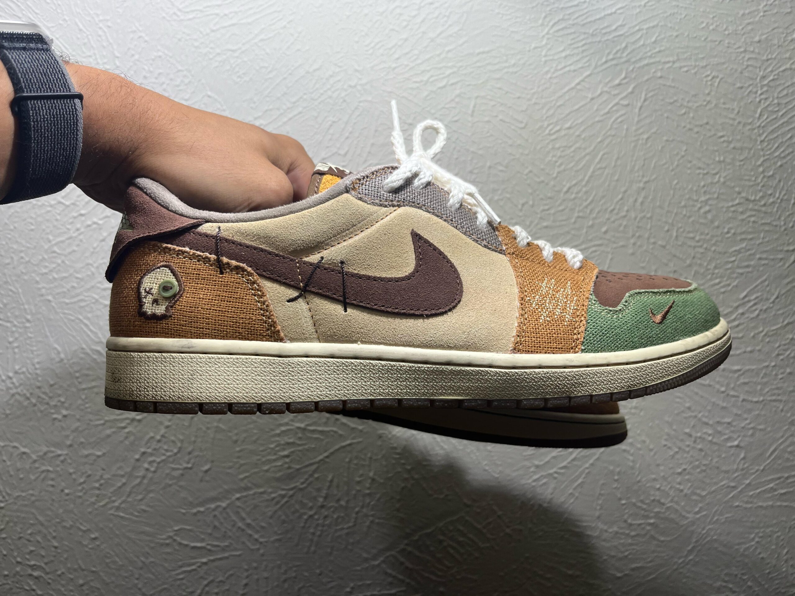 A hand holds a multicolored Nike sneaker with brown, tan, green, and orange panels, white laces, and a stitched Swoosh logo. A small skull emblem is visible near the heel. The background is a textured white wall.