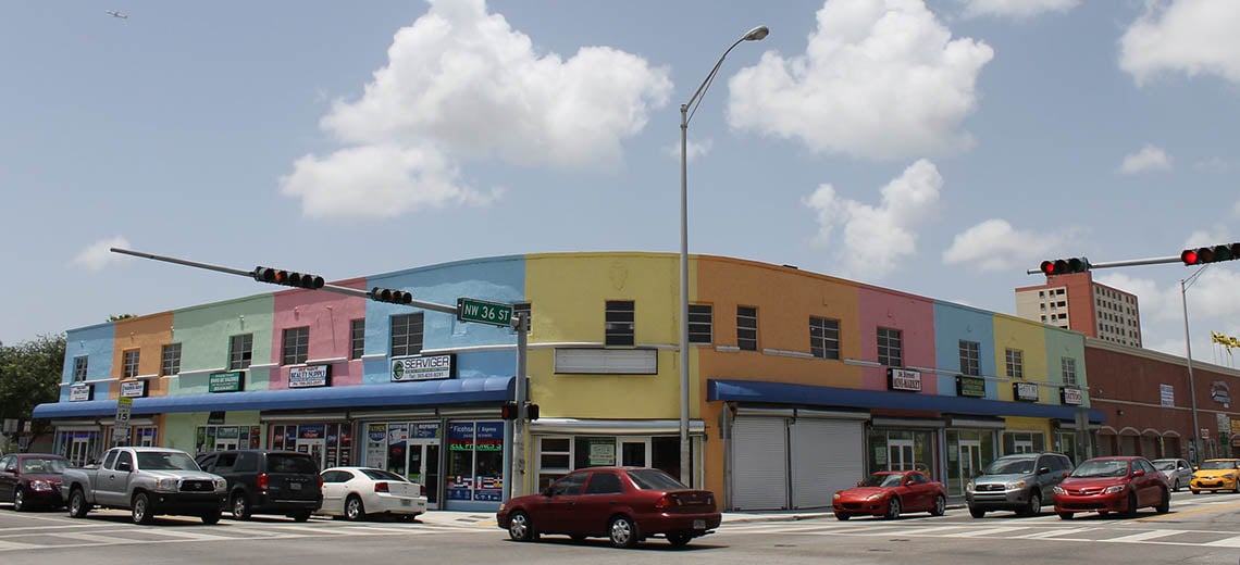 A two-story building with pastel-colored walls houses various shops on a busy street corner. Several cars are stopped at the intersection under a partly cloudy sky. Traffic lights hang overhead.