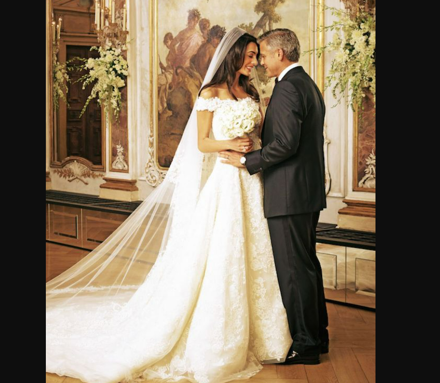 A bride in an off-shoulder white gown and veil holds a bouquet while smiling at a groom in a black suit. They stand closely together in an elegant, ornate room with large floral arrangements.
